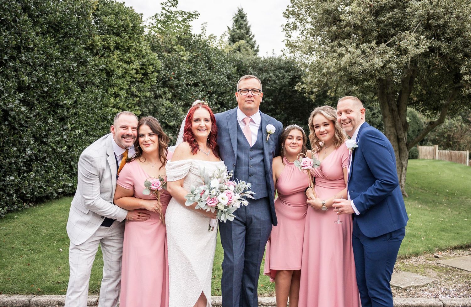 Wedding party in pink and blue attire poses for formal group photo in garden setting.
