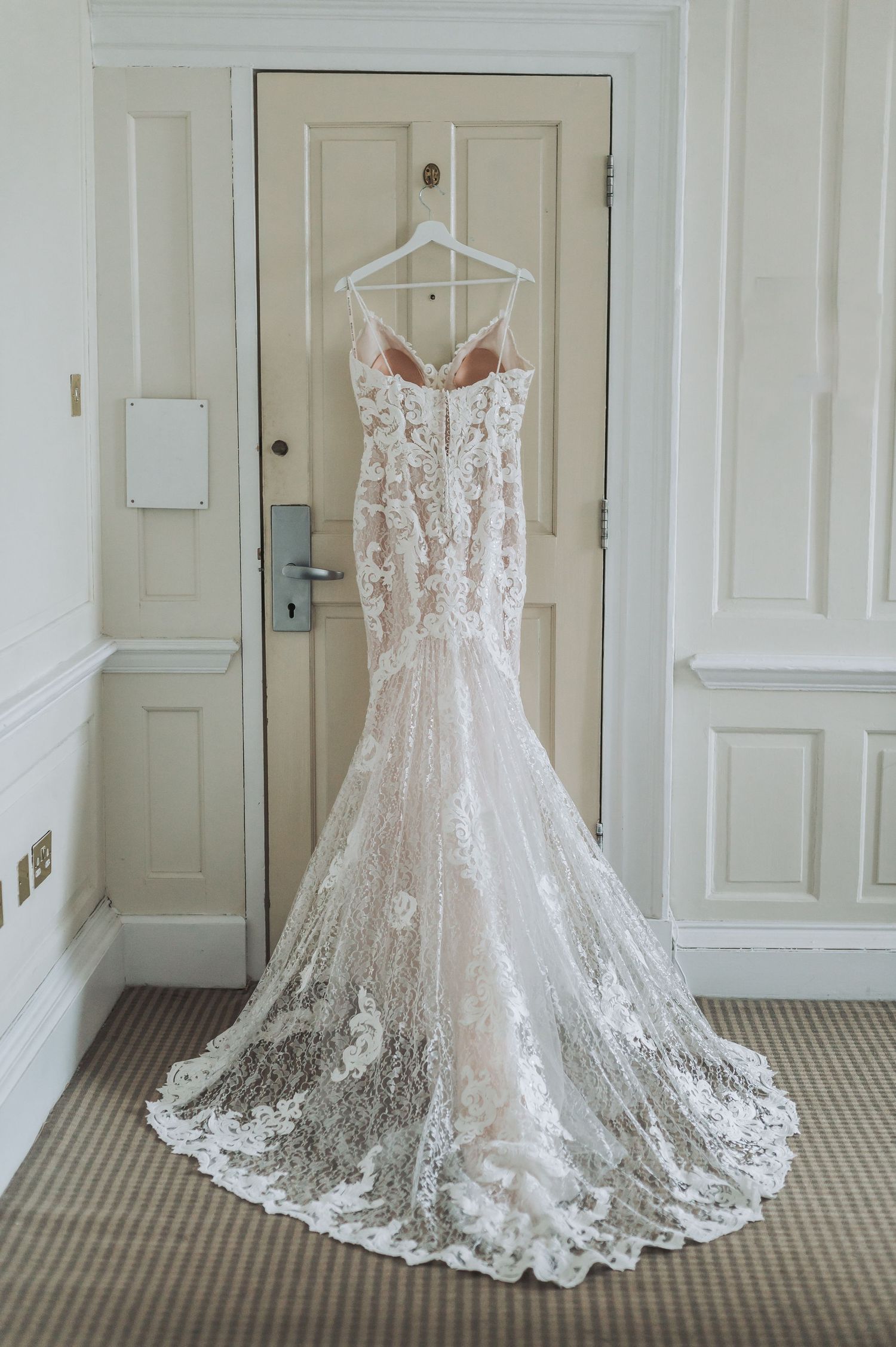 A stunning lace mermaid wedding dress hangs on display against a white door.