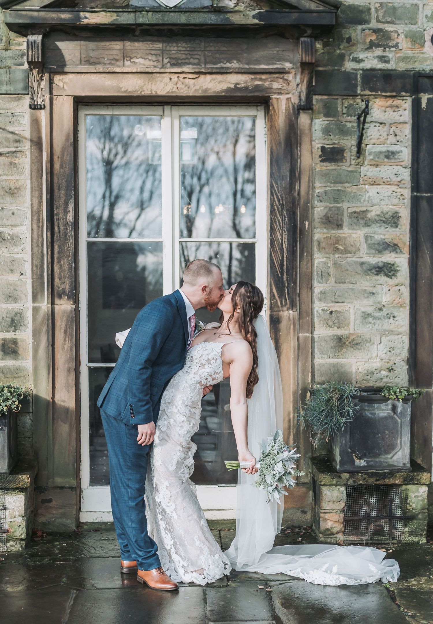 A romantic wedding moment against a rustic stone building with a lace dress and navy suit.