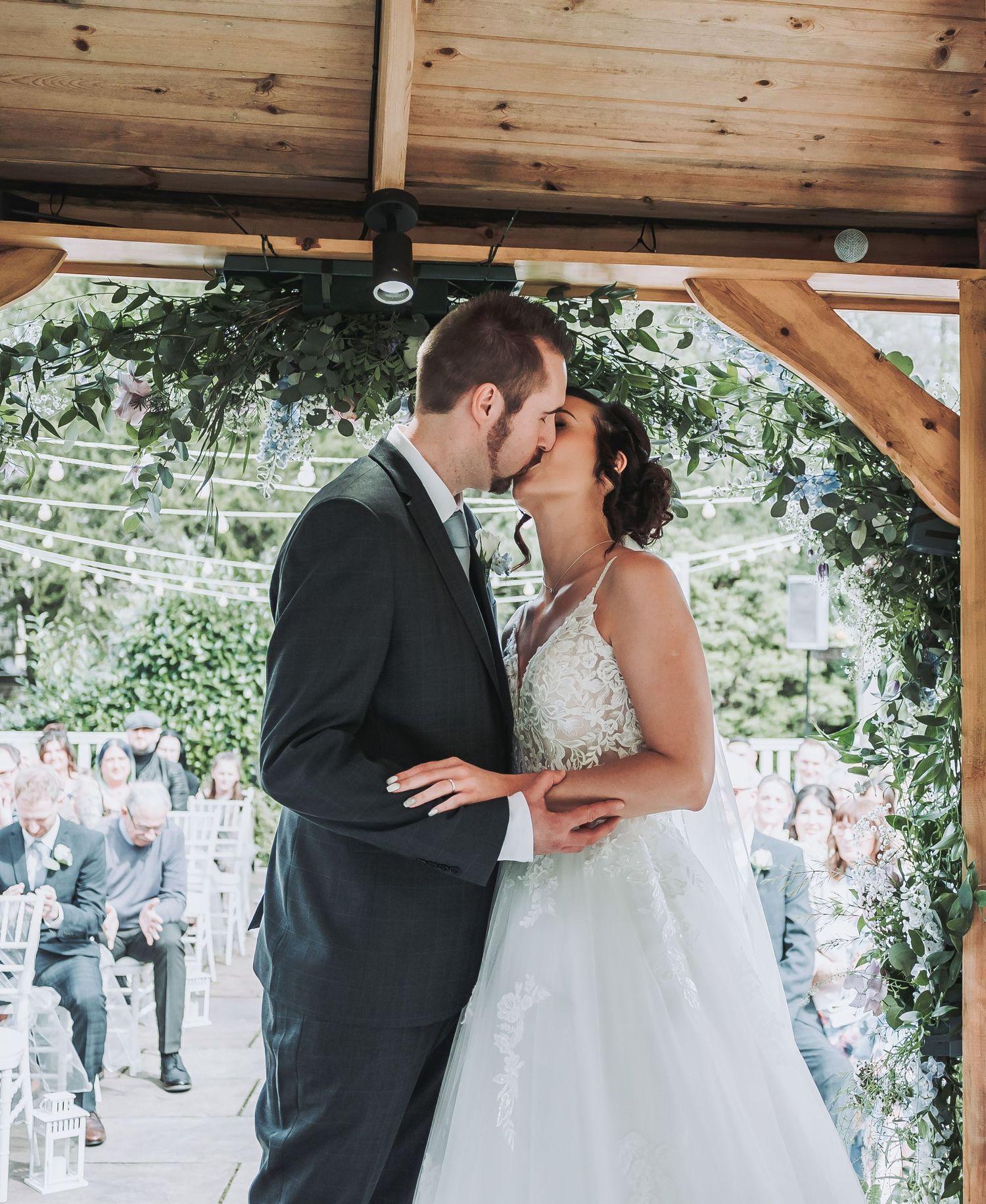 A tender kiss shared between newlyweds under a greenery-adorned wooden pergola.