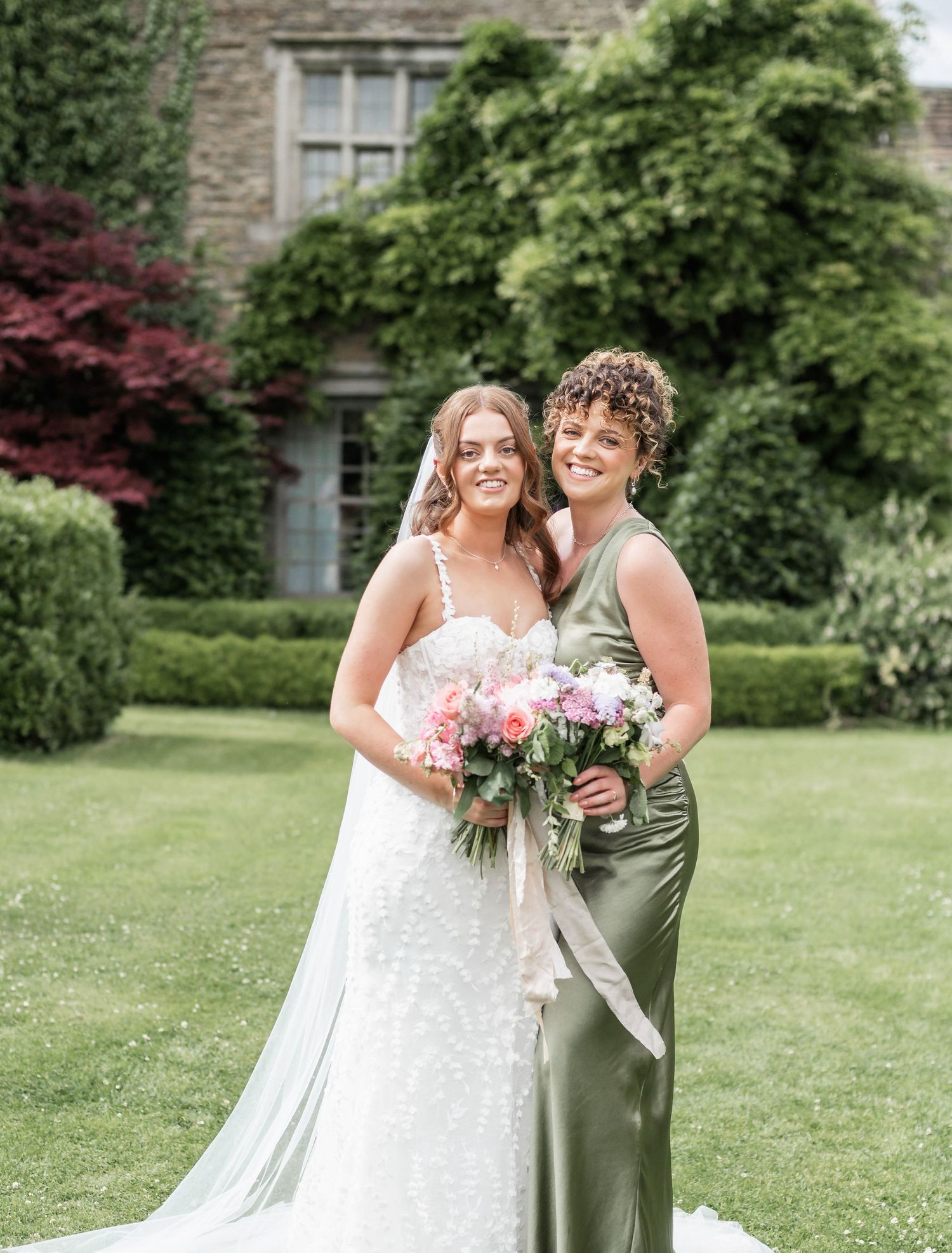 Bride in white gown poses with bridesmaid in sage green dress holding matching bouquets.