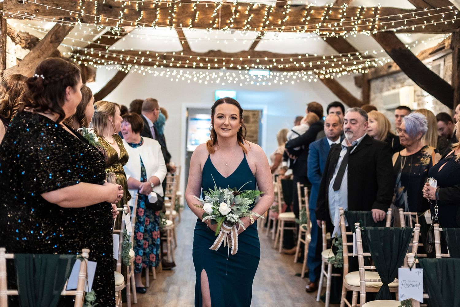 Wedding attendant in navy dress walking down aisle decorated with string lights holding bouquet.