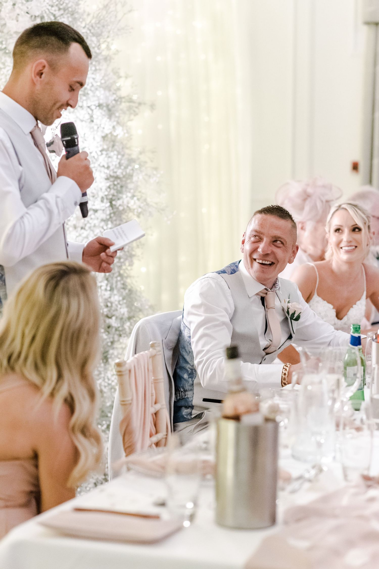 Wedding reception scene with guests enjoying speeches at an elegantly decorated table.