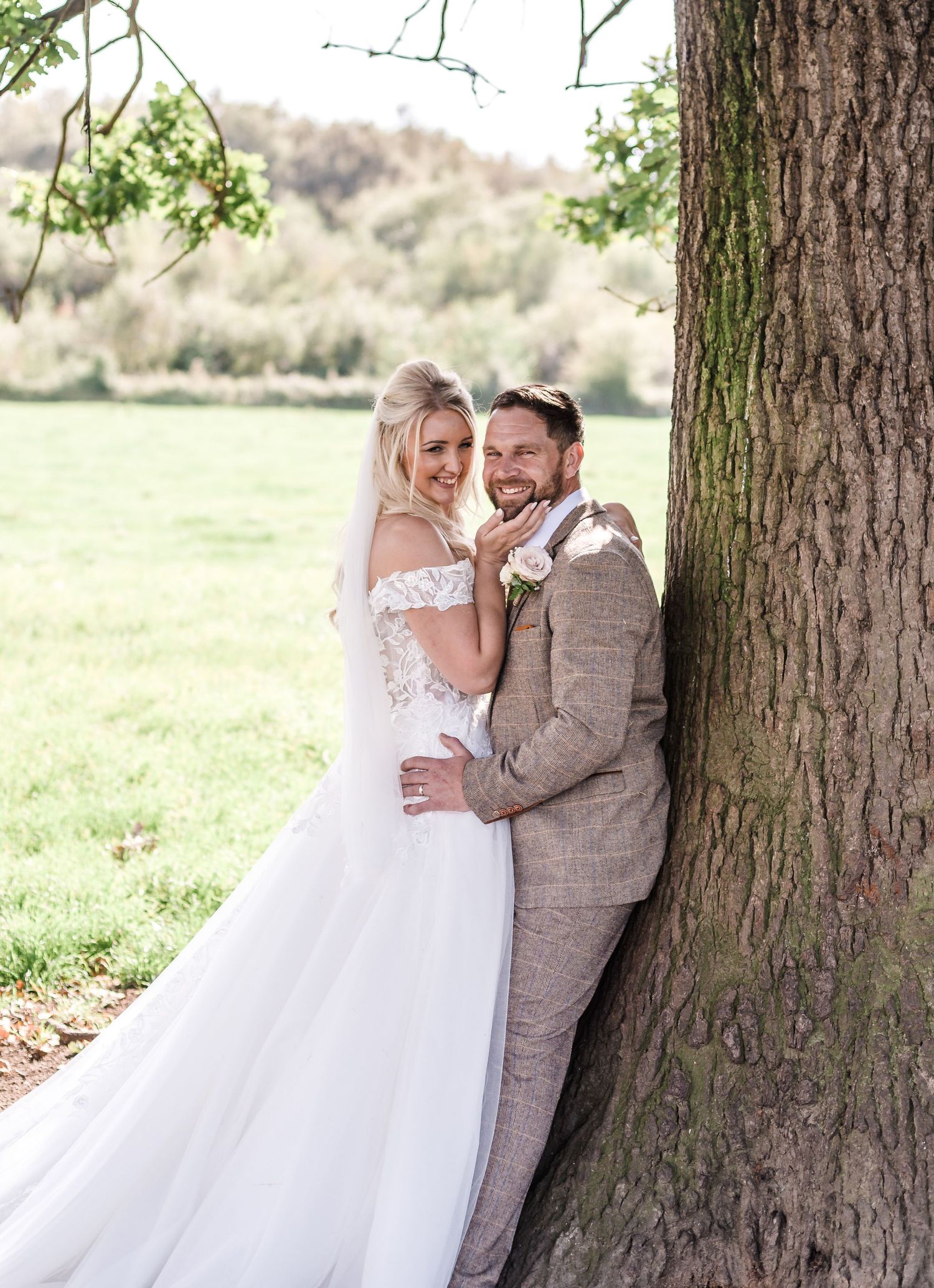 Romantic wedding couple embracing against a large tree trunk in a pastoral setting.