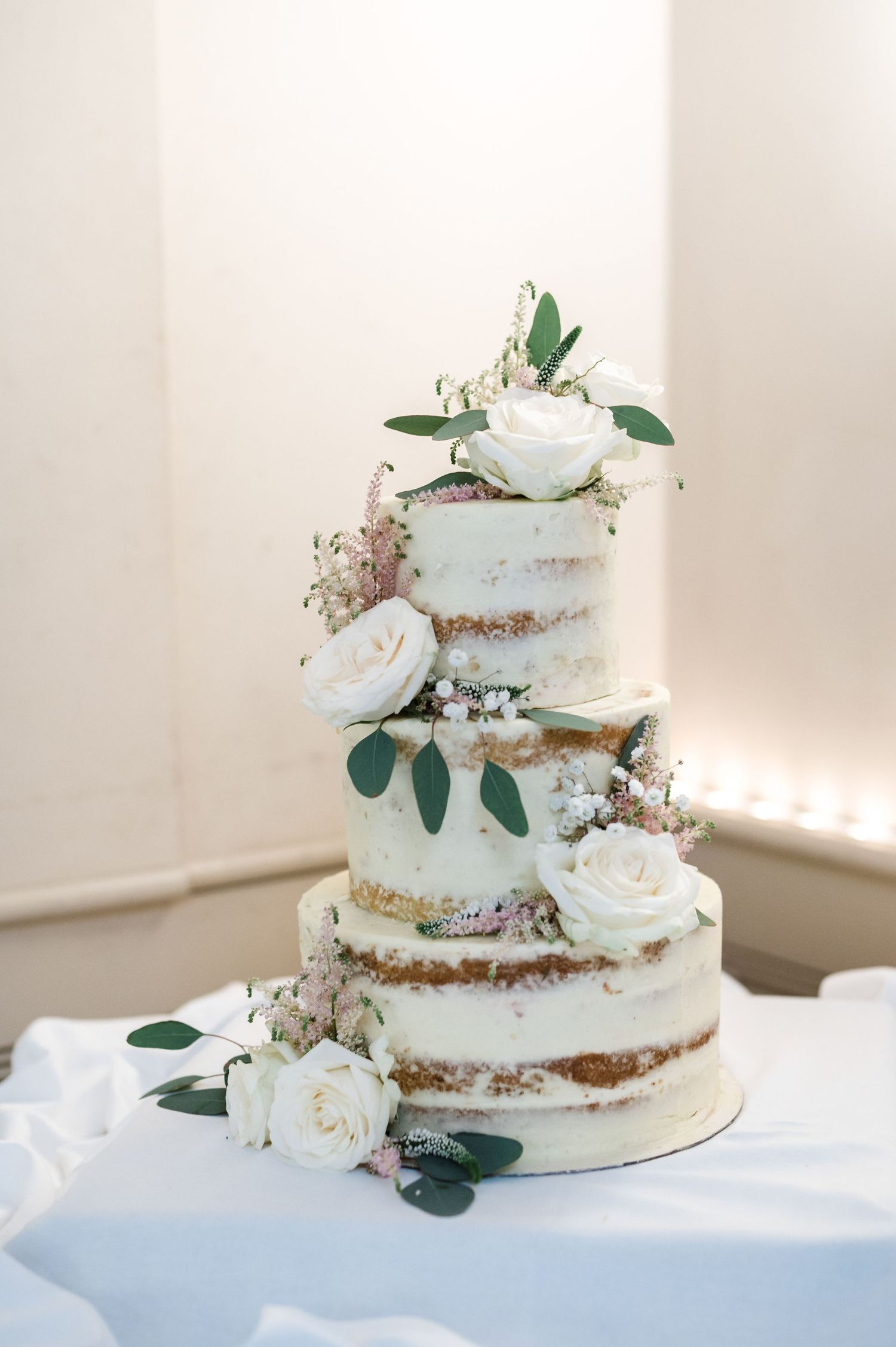 Semi-naked wedding cake decorated with white roses and eucalyptus leaves on a white tablecloth.