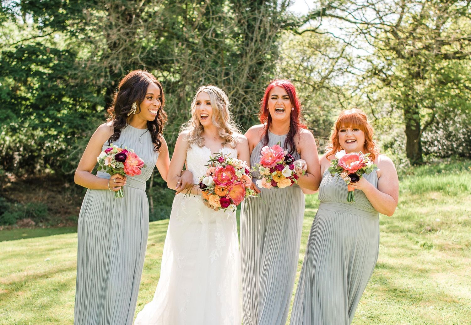 Wedding party holding colorful floral bouquets poses together on sunny grass lawn.