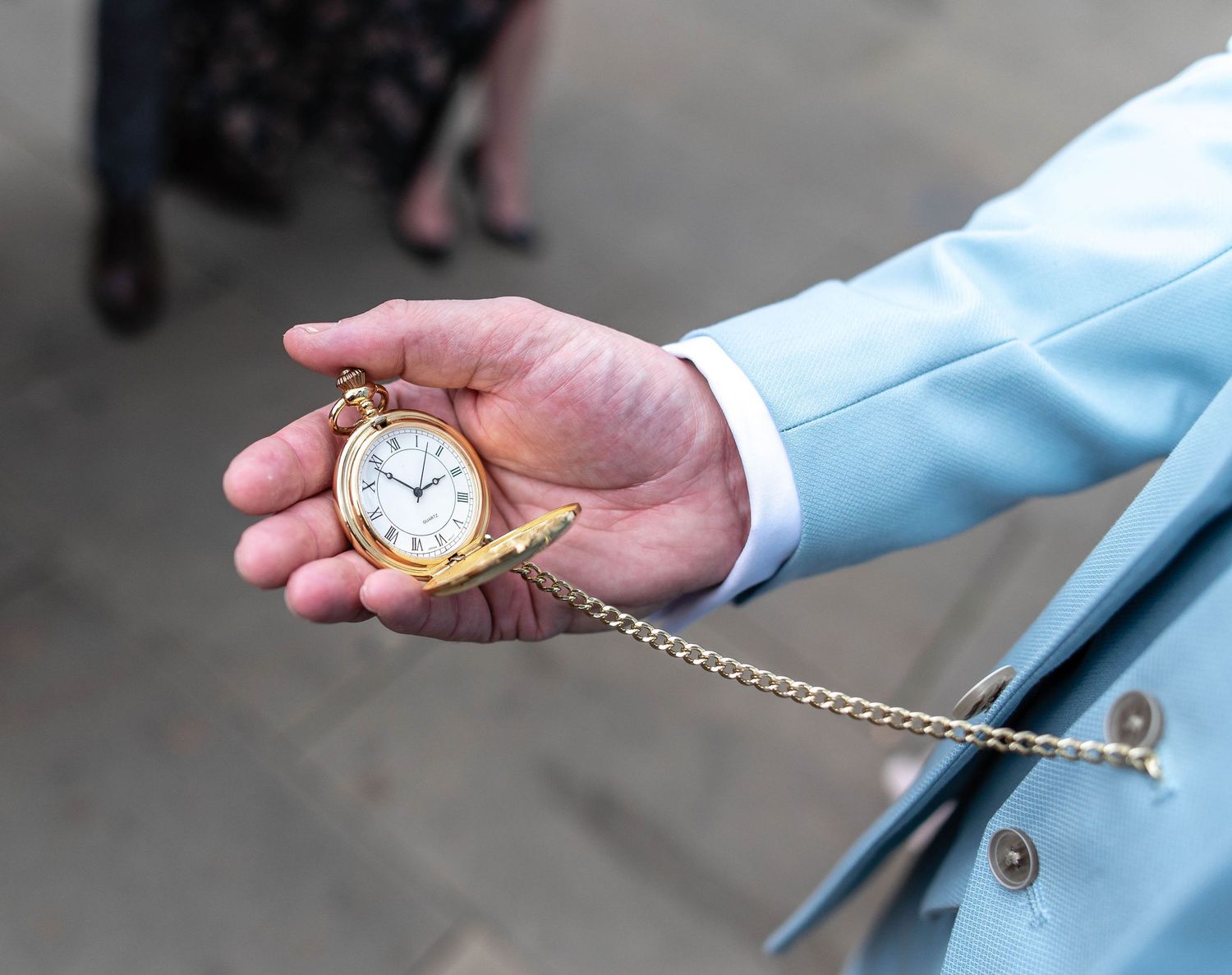 Vintage gold pocket watch on chain held in hand wearing light blue suit sleeve.