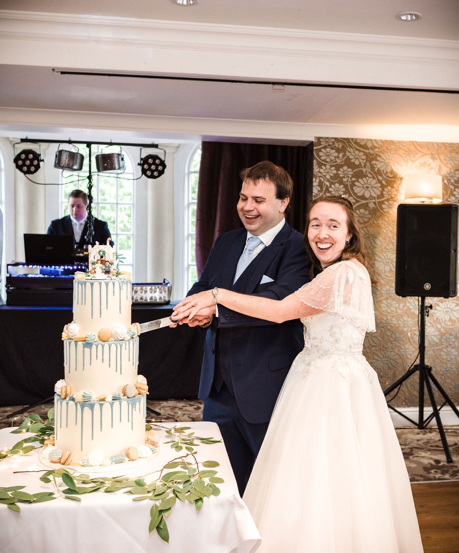 Newlyweds cut into an elegant three-tiered white wedding cake decorated with greenery.