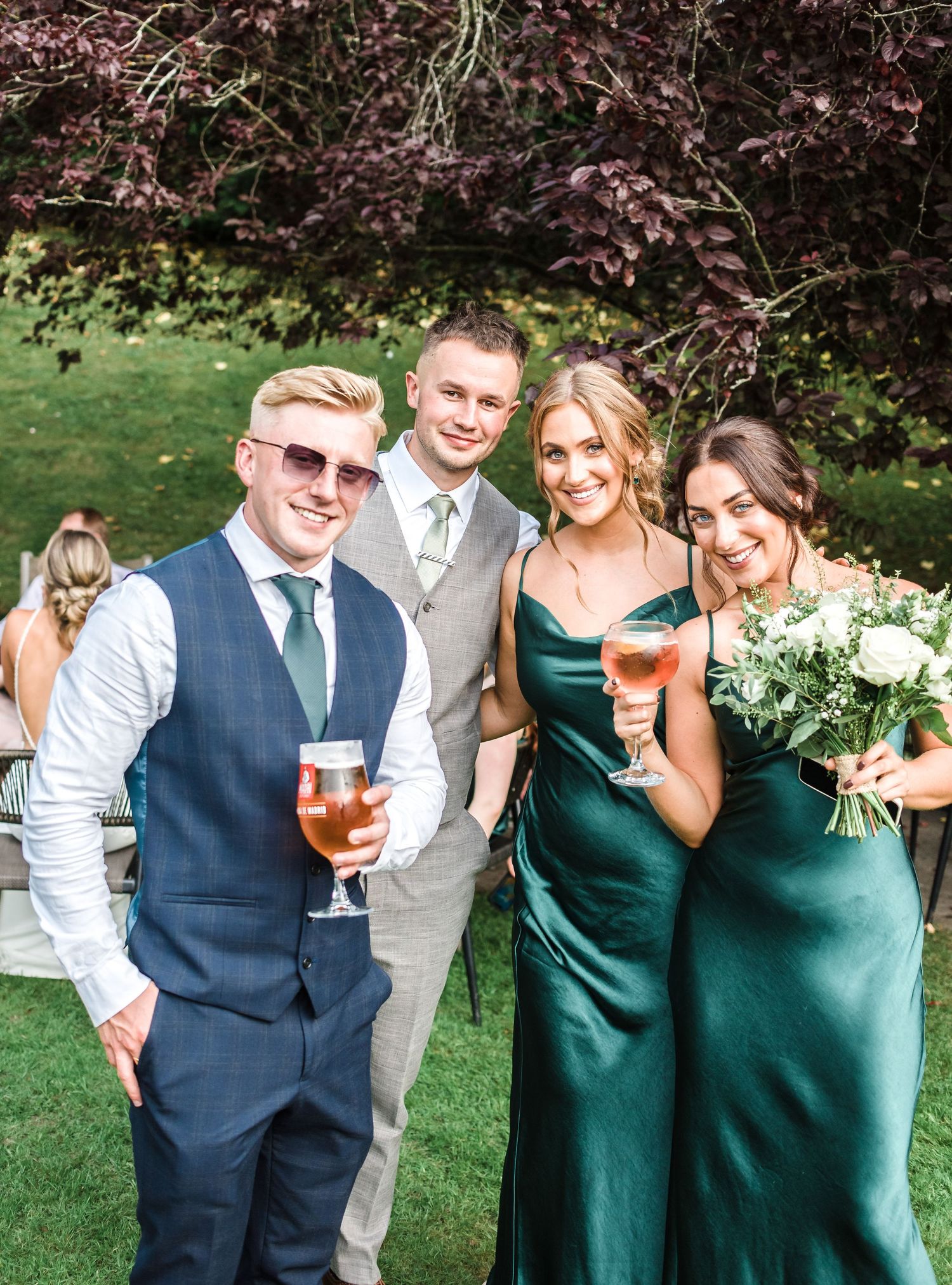 Wedding party members in green attire and navy vests enjoying drinks outdoors.