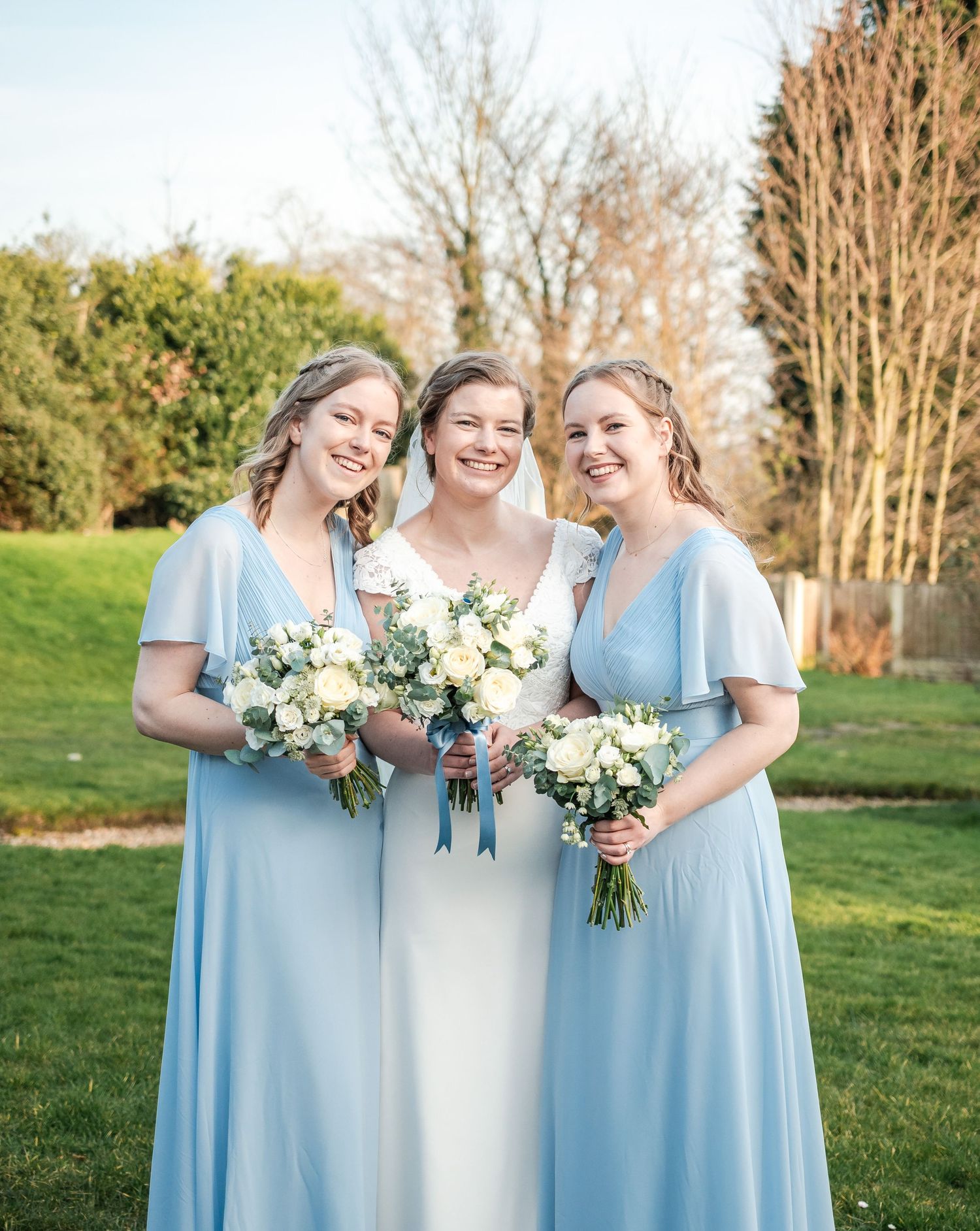 Three women in pale blue dresses holding white bouquets pose together at an outdoor wedding.