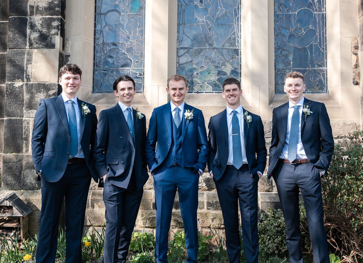 Groomsmen in navy blue suits pose together against a stone wall with greenery in an outdoor wedding setting.