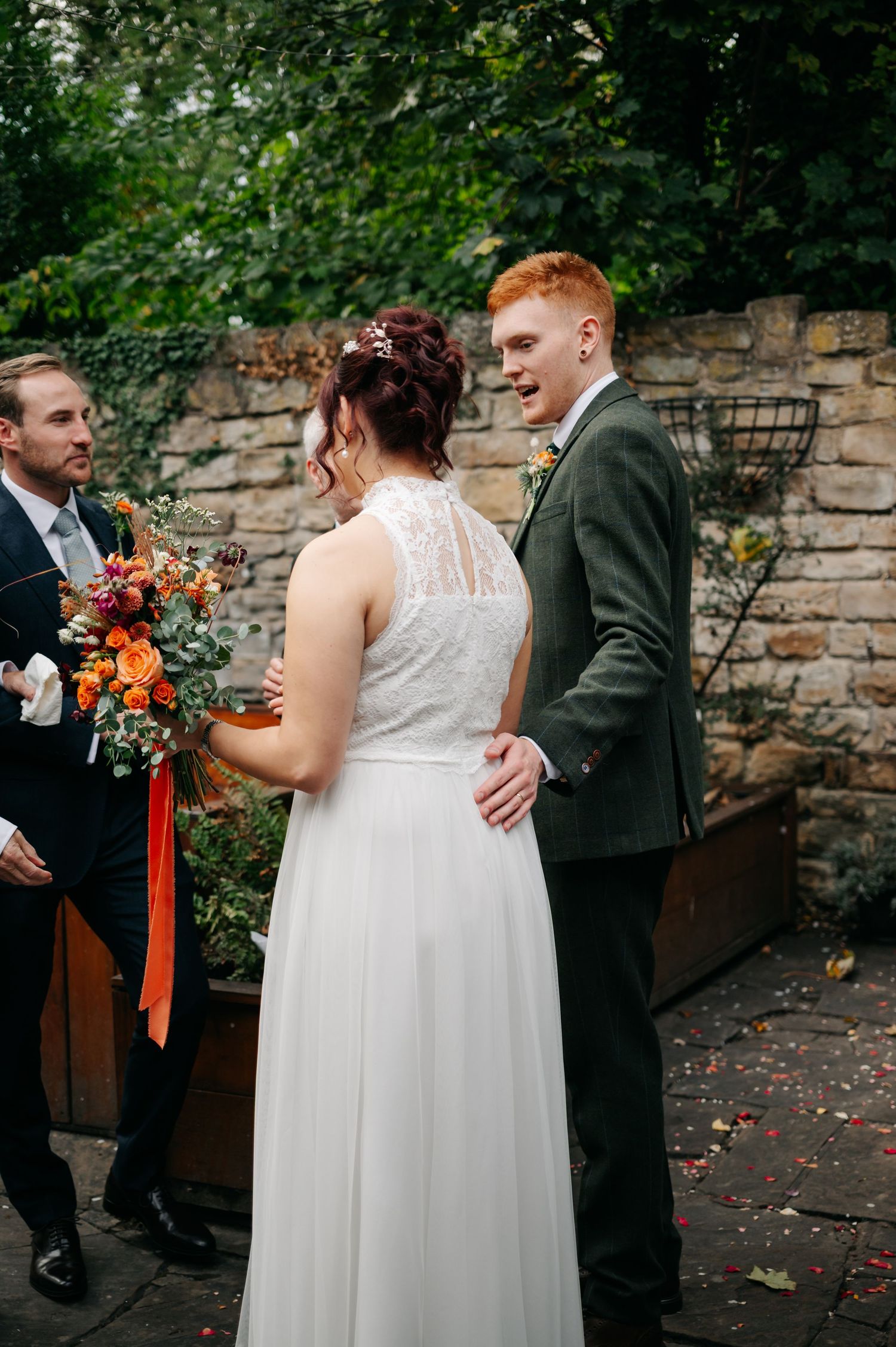 Wedding couple and guests gather outdoors near stone wall with lush greenery in background.