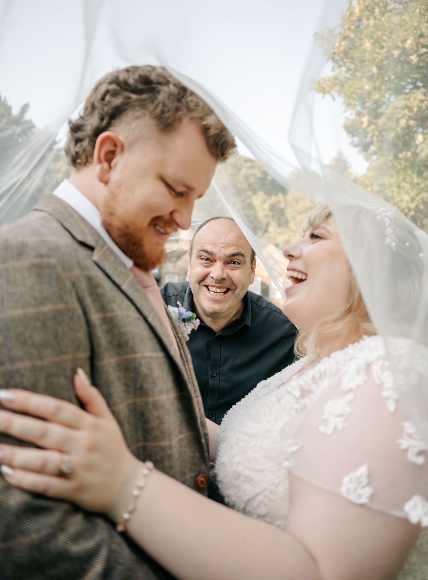 Bride and groom share a joyful moment under a veil as a smiling officiant looks on at their wedding.