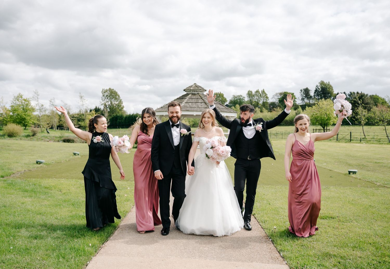 Wedding party celebrating outdoors on a garden path, bride in white gown with bridesmaids in pink and black dresses.