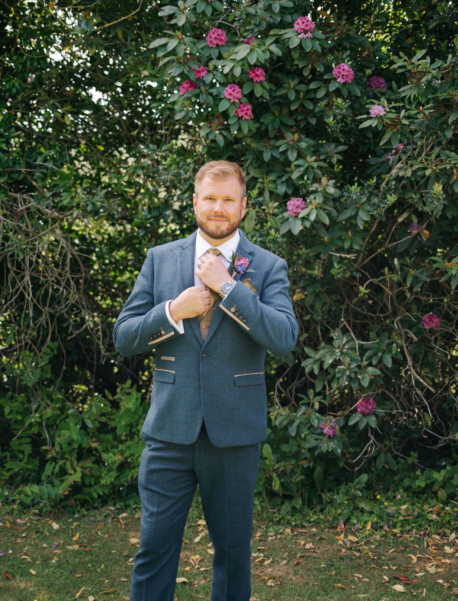 Groom in a navy suit poses for portraits near flowering pink bushes in a garden setting.