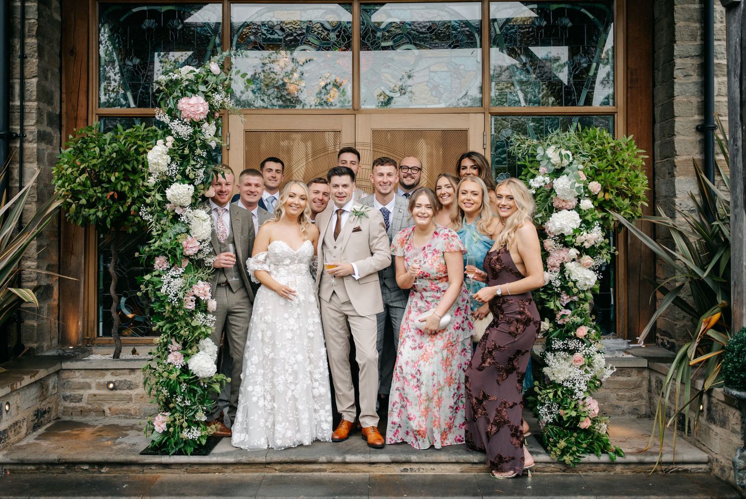 Wedding group photo taken in front of ornate wooden doorway with floral arch decorations.
