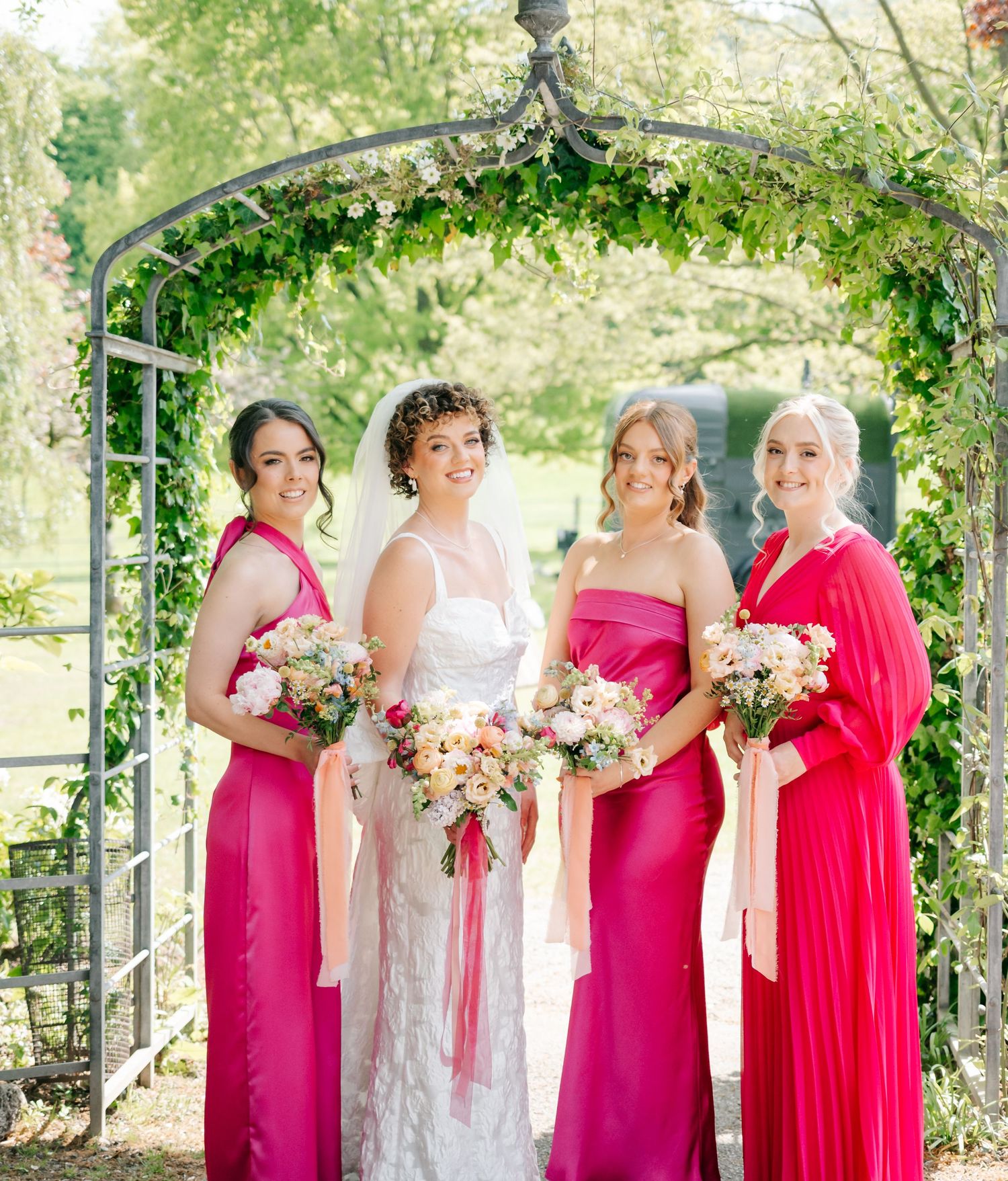 Bridesmaids in vibrant pink dresses stand with bride under an outdoor garden arch.