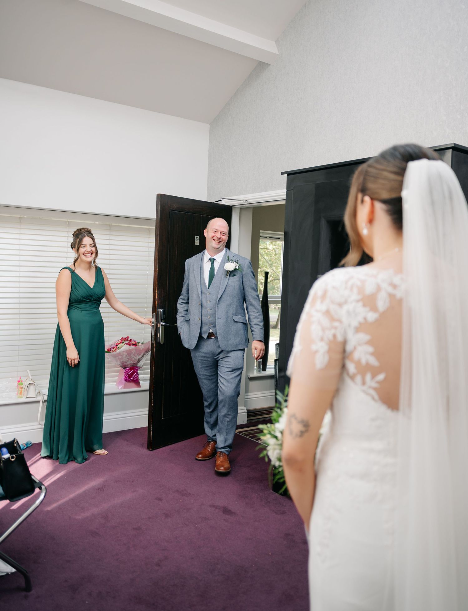 First look moment captured in a room with emerald bridesmaid dress and gray suit visible.
