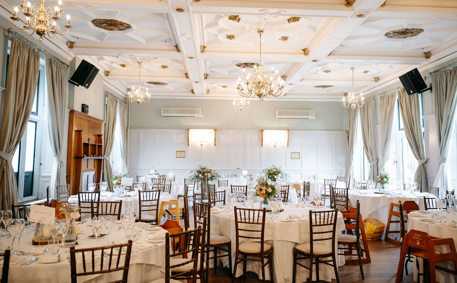 Elegant wedding reception room with round tables, wooden chairs, crystal chandeliers, ornate coffered ceiling and tall draped windows.