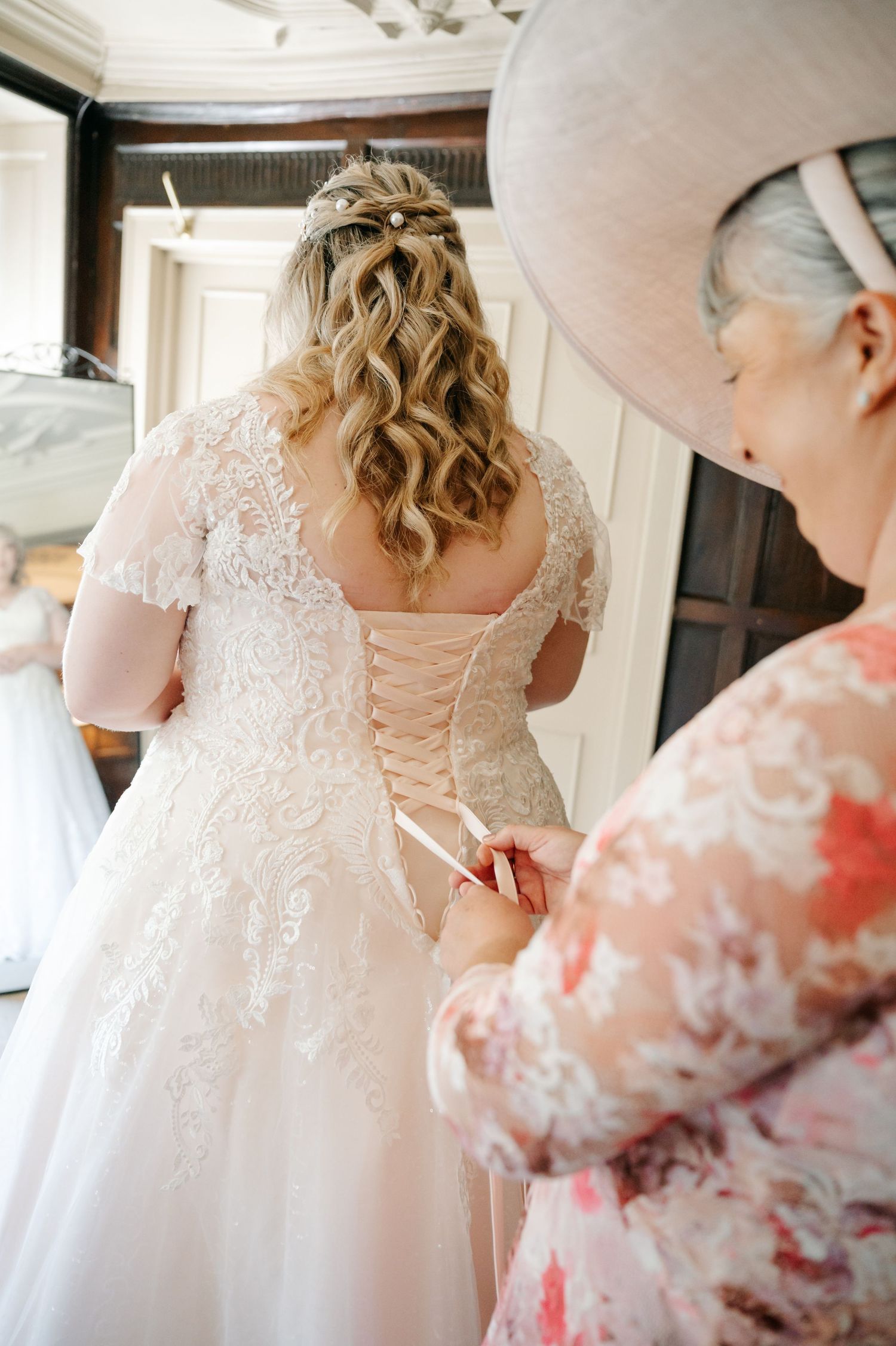Bride in lace wedding dress being laced up by older woman in floral dress and hat.