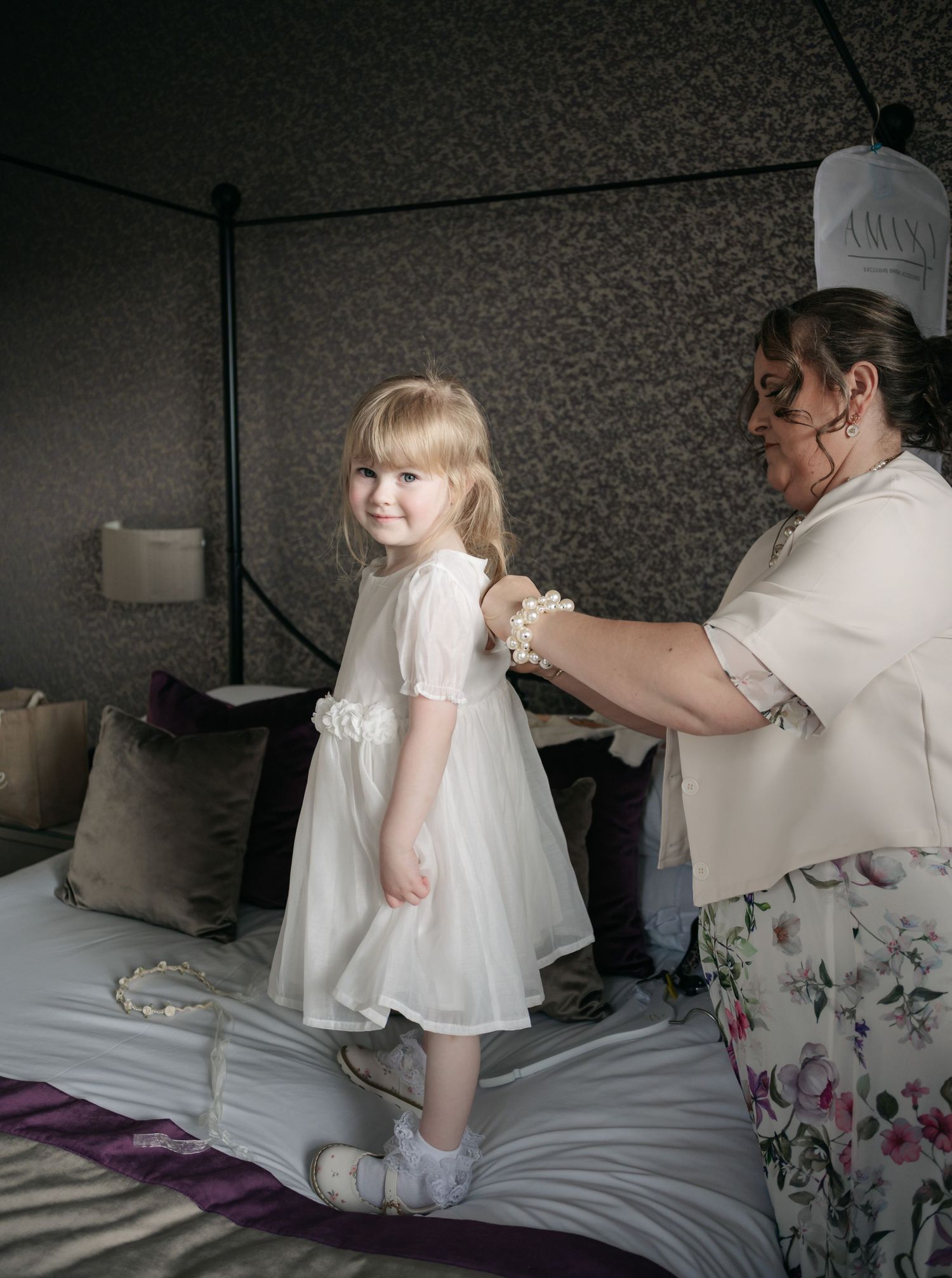 A young child in a white dress plays on a bed during wedding preparations.
