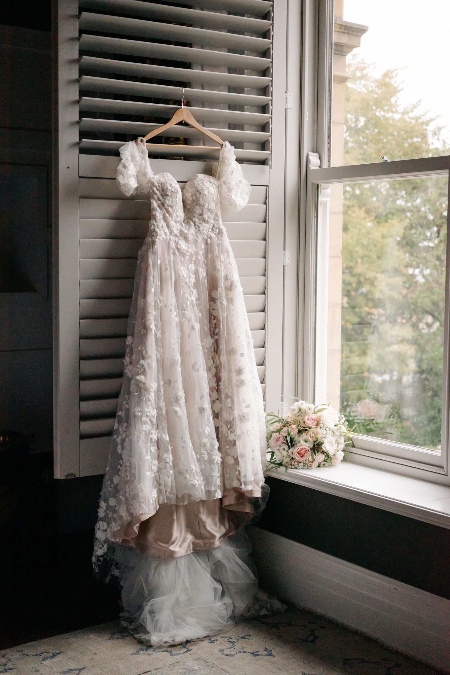 A delicate white lace wedding dress hangs by a window, showcasing intricate details and flowing fabric.