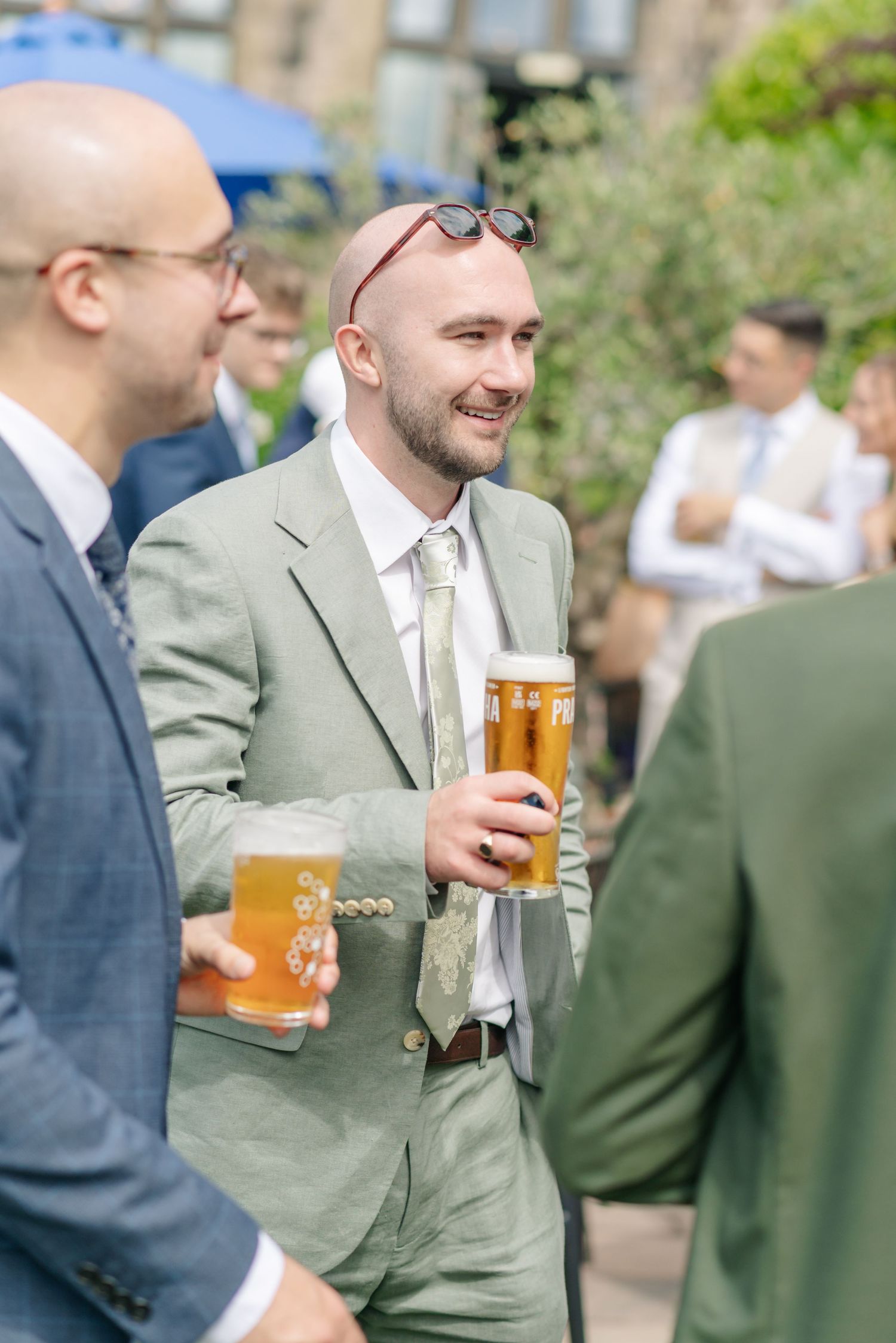 Wedding guests in suits enjoying beers and conversation in an outdoor garden setting.