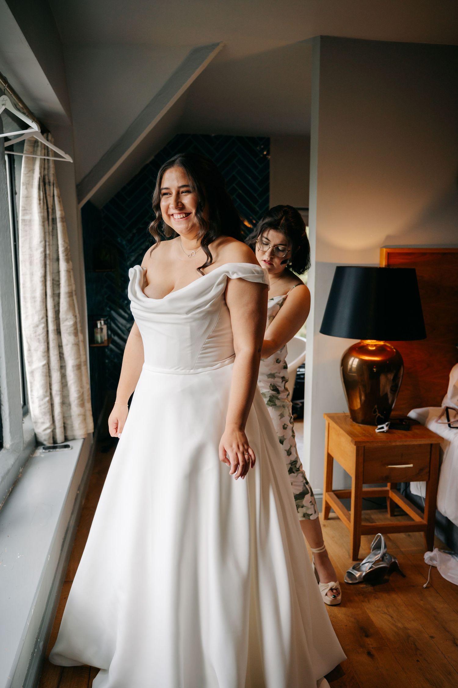 Getting ready scene showing a white wedding dress being put on in a warmly lit room.