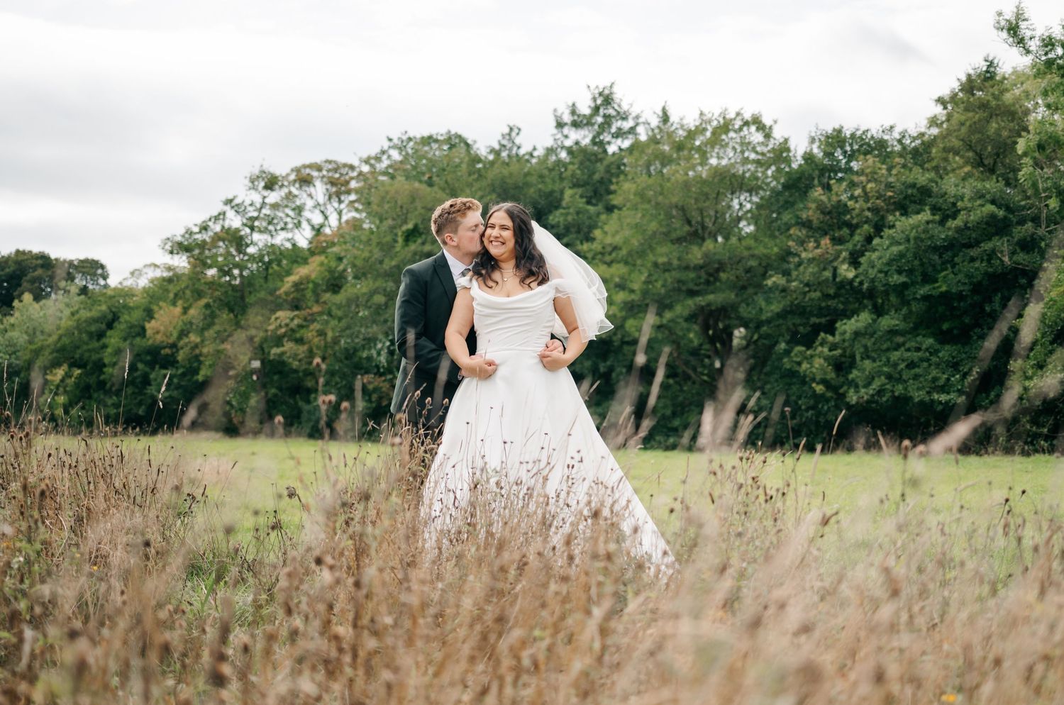 A couple in wedding attire embrace in a rustic field with tall grass and trees in the background.