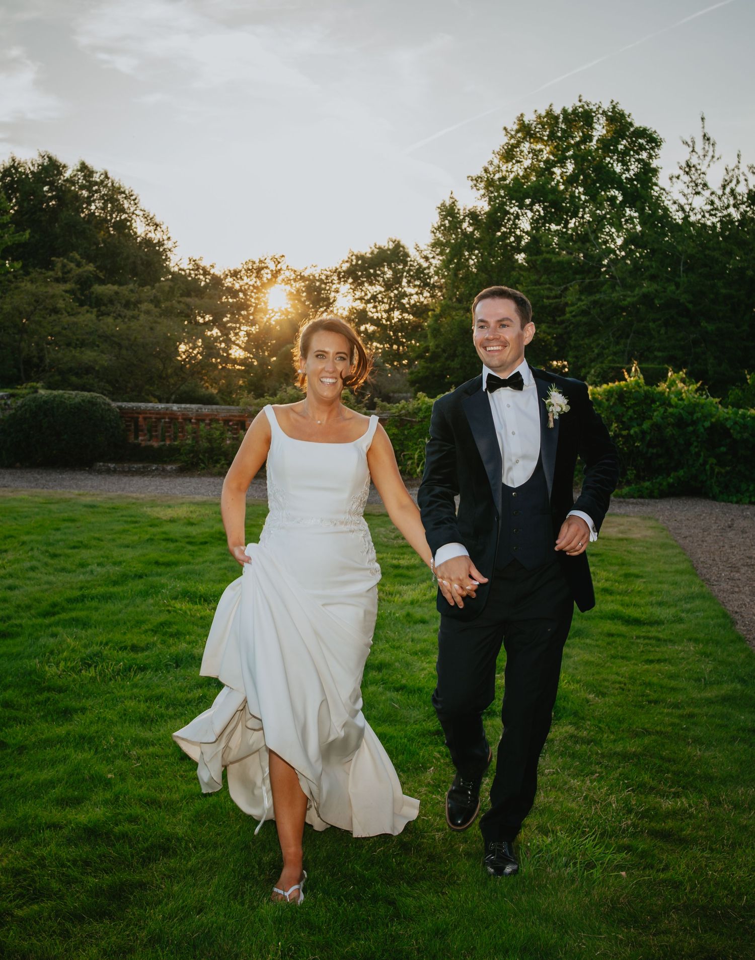 Joyful couple runs through garden path at sunset during wedding celebration.