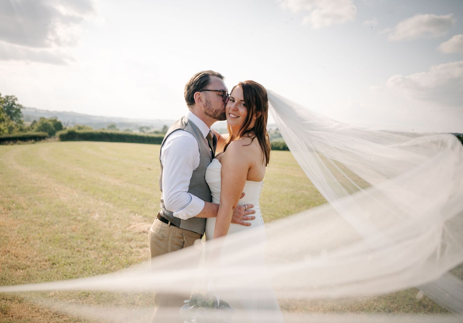 Bride and groom share a romantic kiss outdoors on their wedding day, with a flowing veil billowing in the breeze.