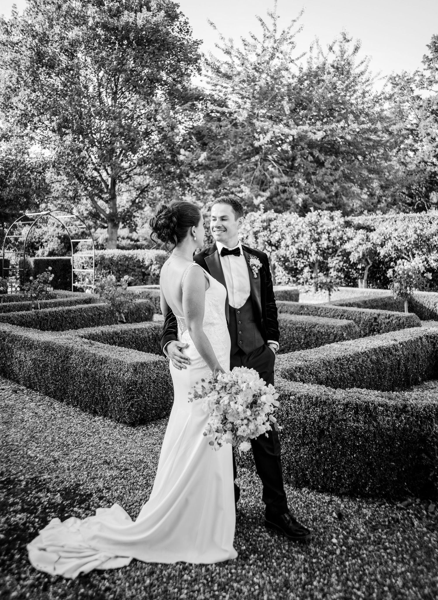 A wedding couple shares a romantic moment in a manicured garden with geometric hedges.