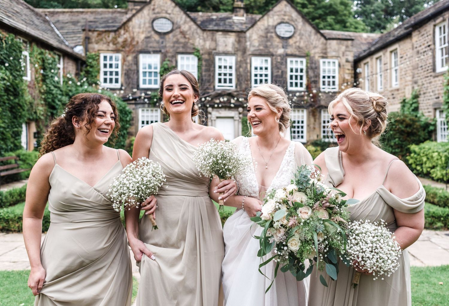 Bridesmaids in neutral dresses hold baby's breath bouquets while laughing together outside a stone building.