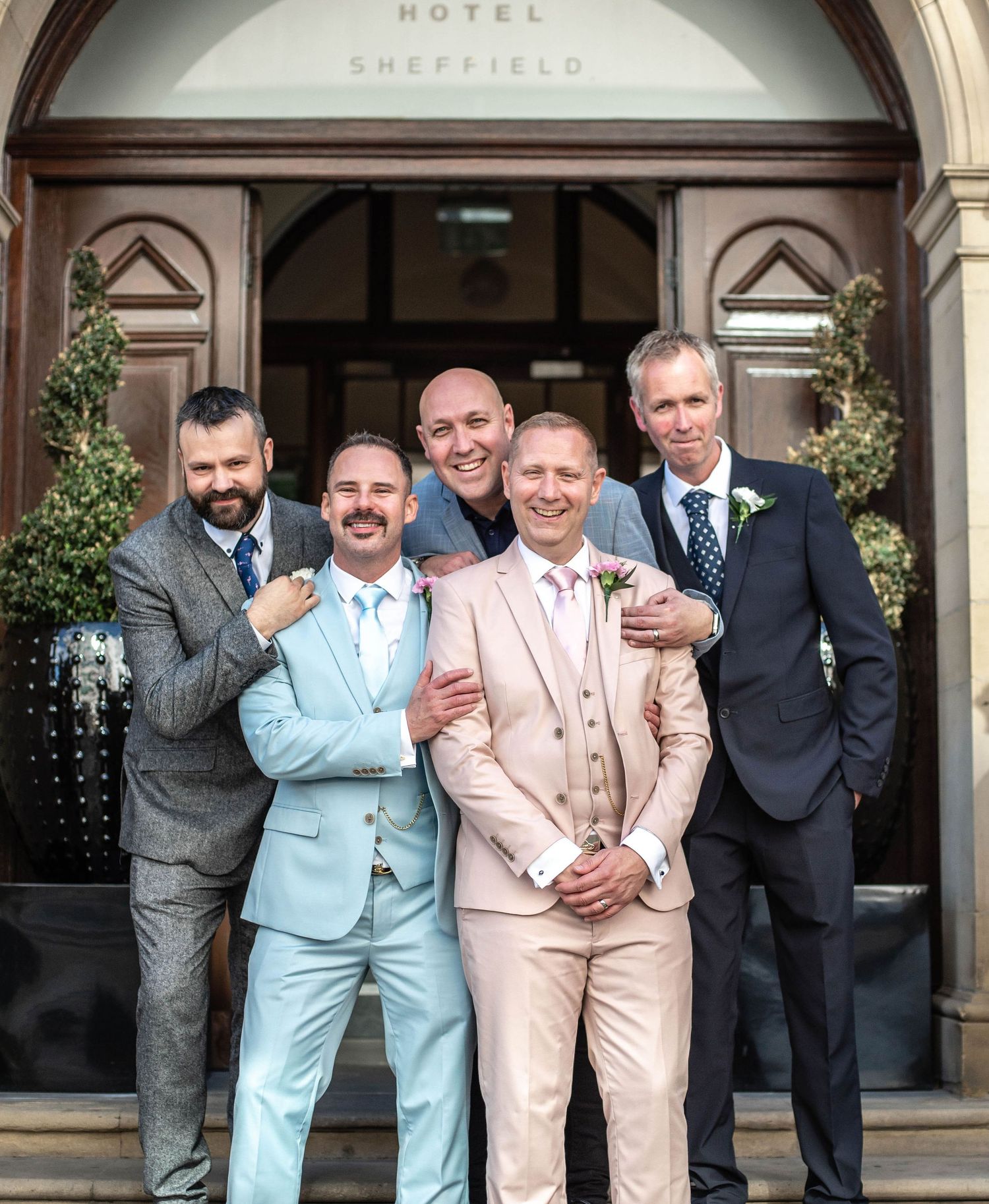 Group portrait of wedding party members in pastel and dark suits outside a hotel entrance.