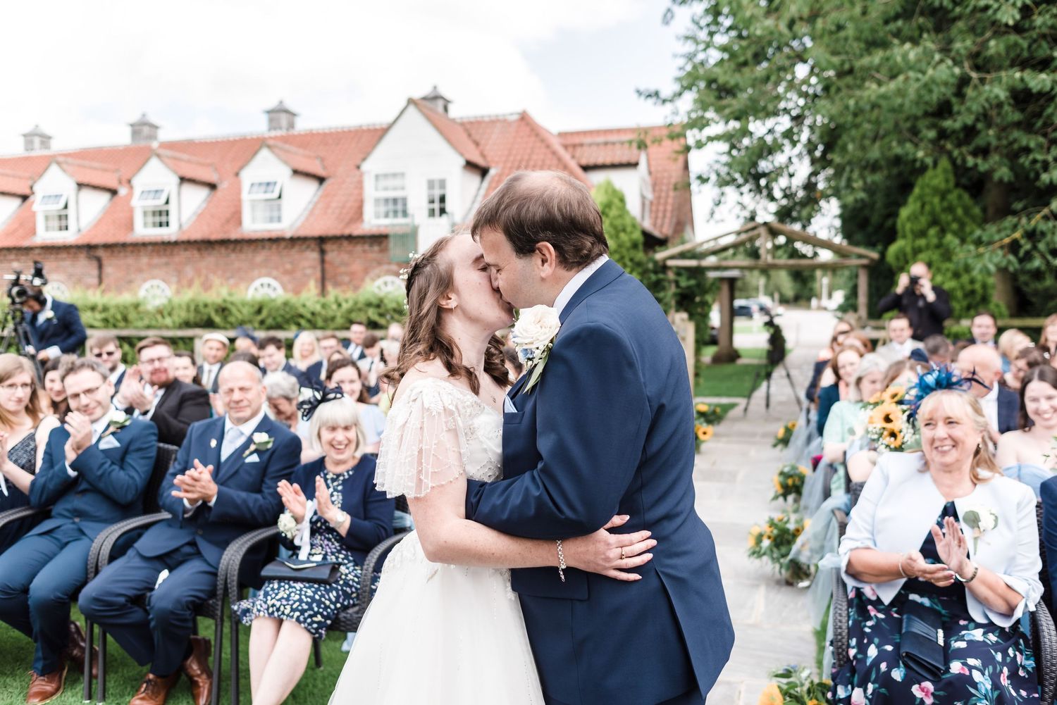 Wedding couple shares a kiss during outdoor ceremony while guests look on from garden seating.