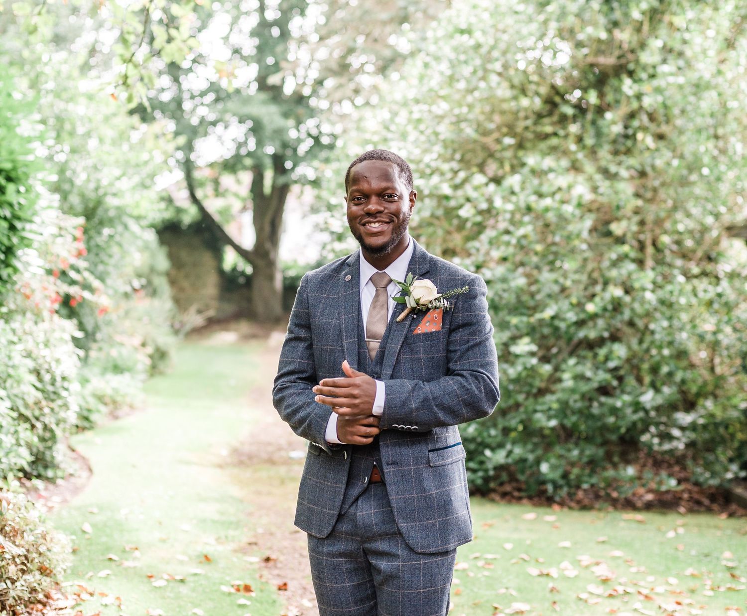 Groom in grey checkered suit with coral boutonniere smiling in garden setting.