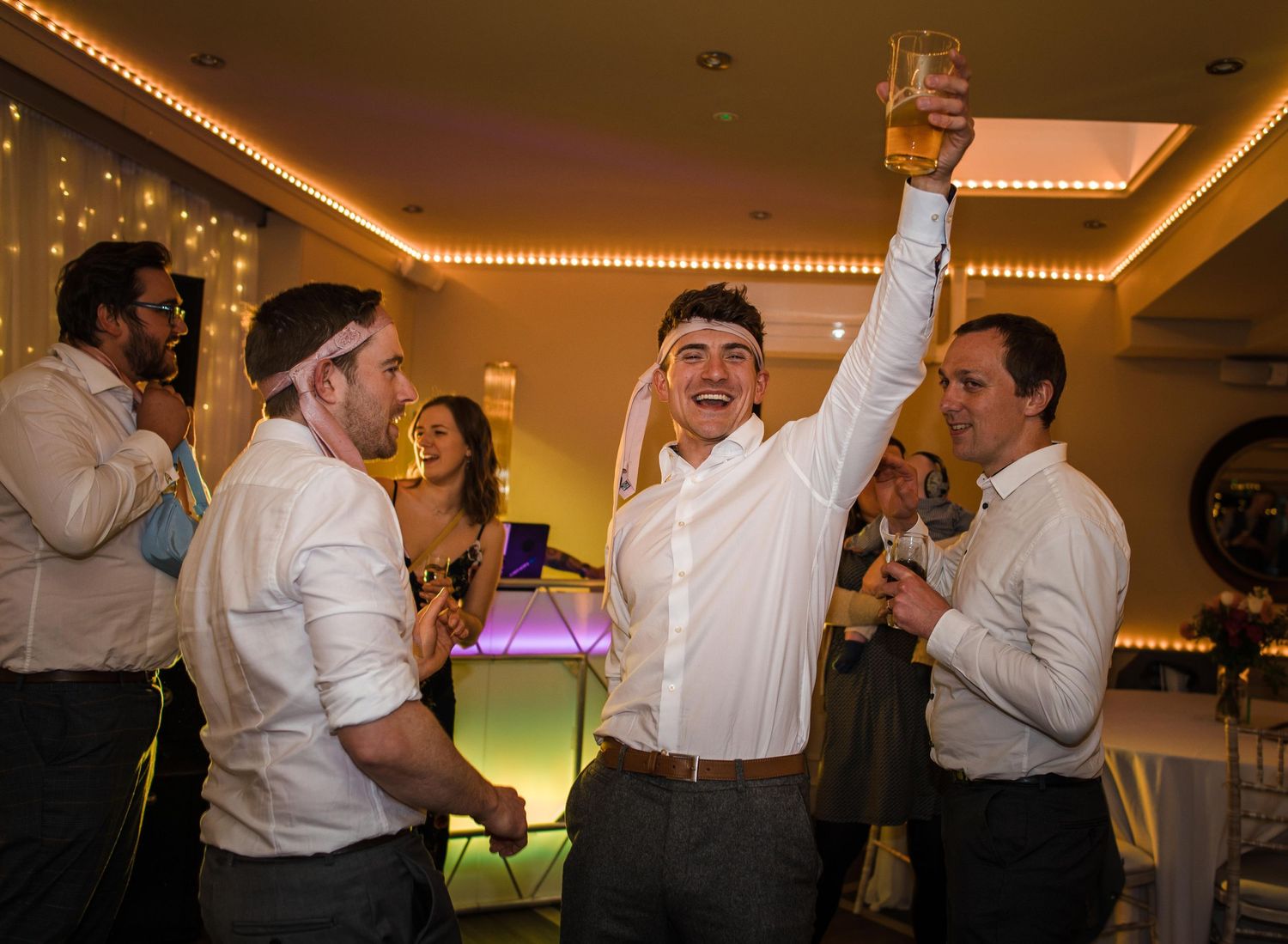 Guests celebrate at a wedding reception with one raising a glass under string lights.