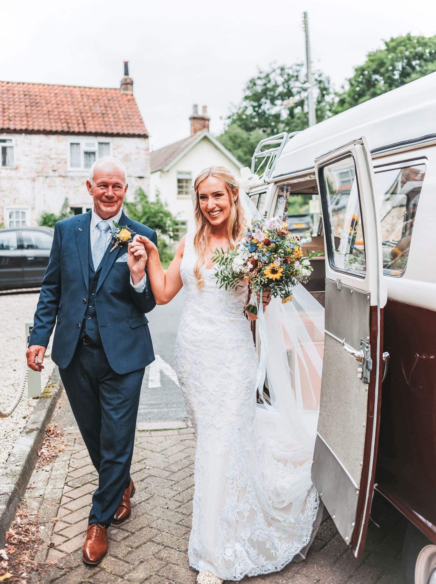 Classic wedding scene with vintage VW van and elegant bridal attire outside stone buildings.