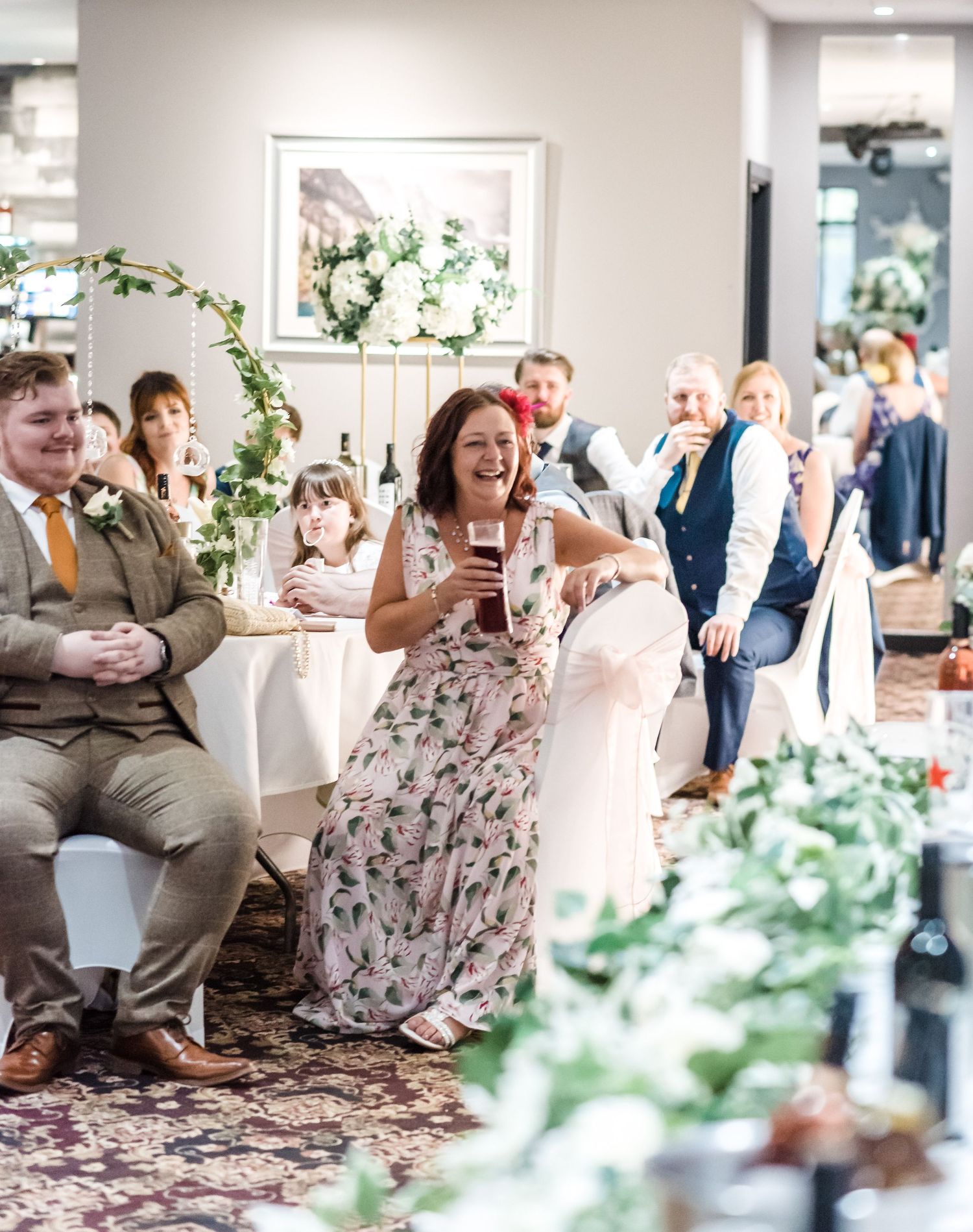 Wedding guests enjoying celebration in decorated reception venue with floral arrangements.