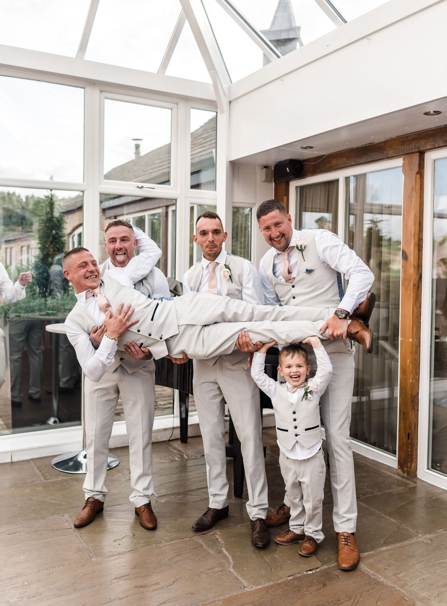 Wedding party in white suits playfully lift a young ring bearer in matching attire.