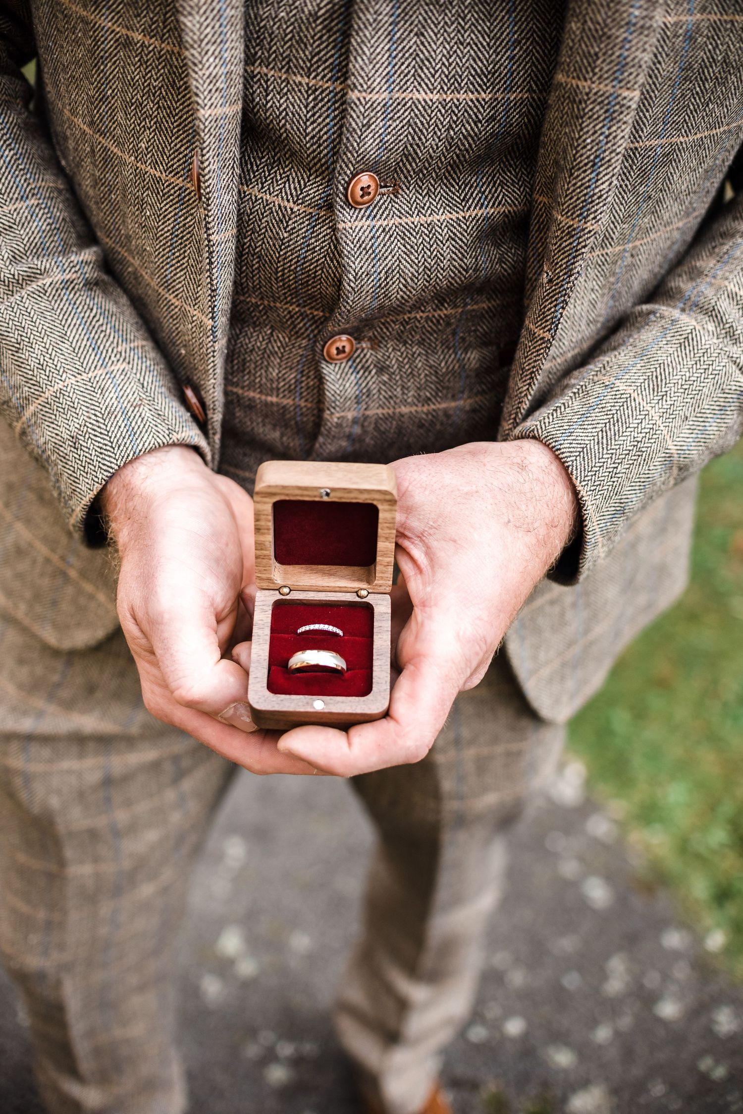 Hands holding a small wooden ring box with wedding band against tweed suit fabric.