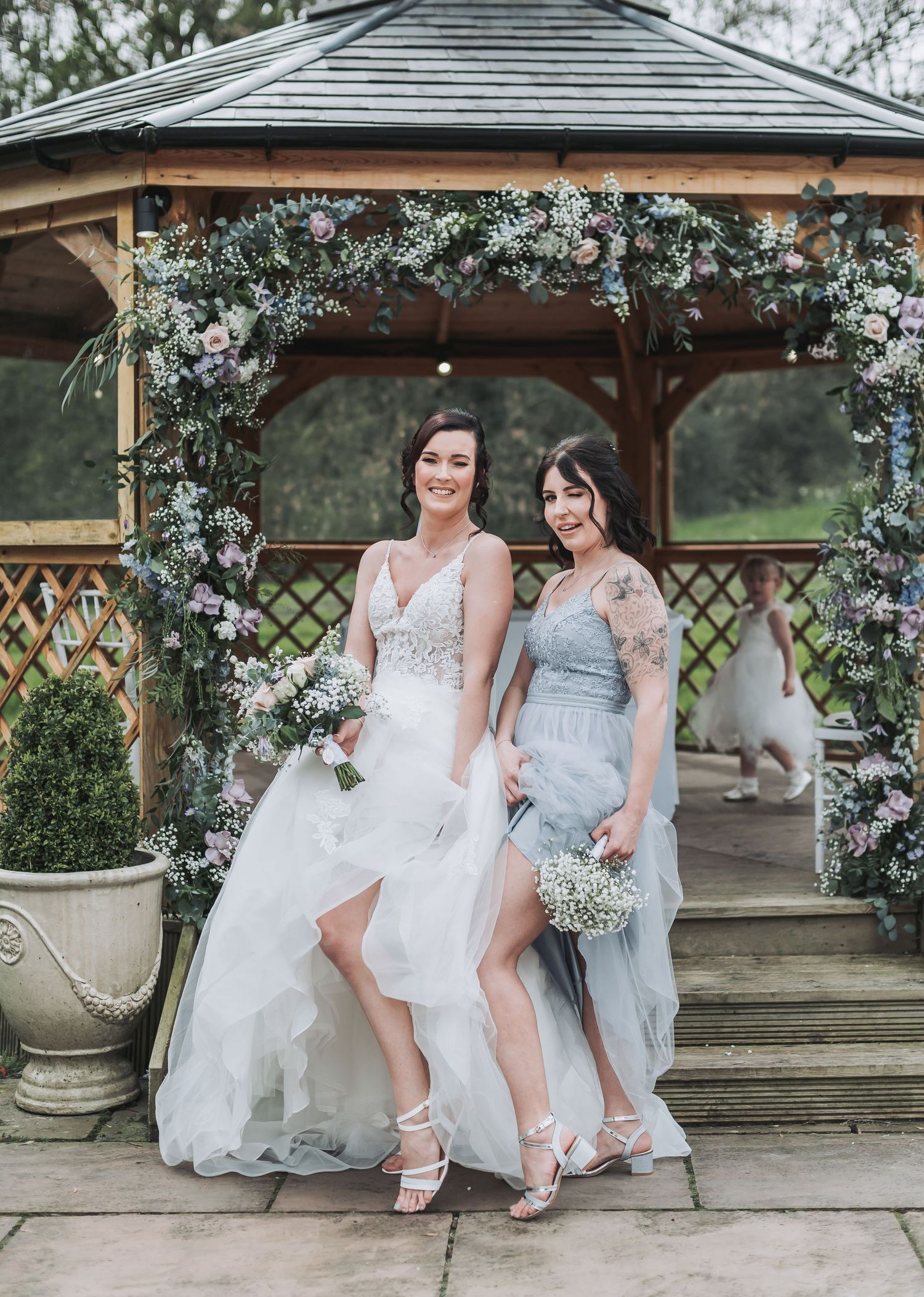 Two people in formal dresses pose together under a gazebo decorated with white flowers.