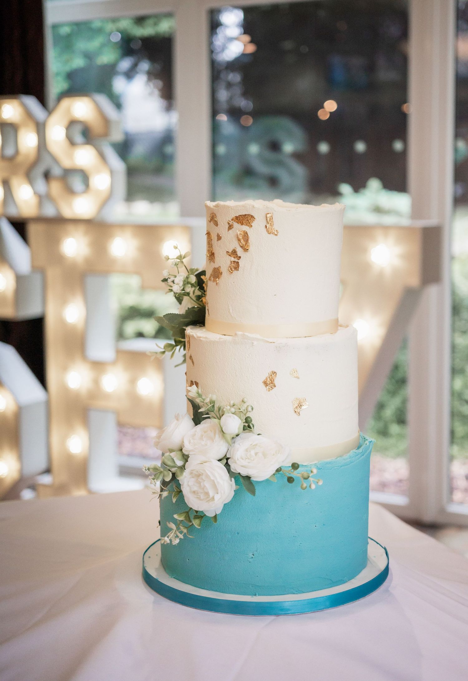 Three-tier wedding cake featuring white and turquoise colors with gold accents and fresh flowers.