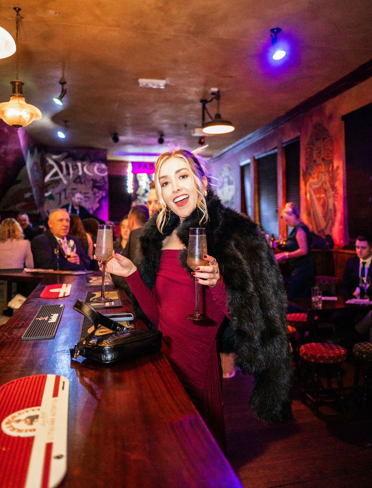 Person in red dress and black fur coat poses at bar with champagne glass.