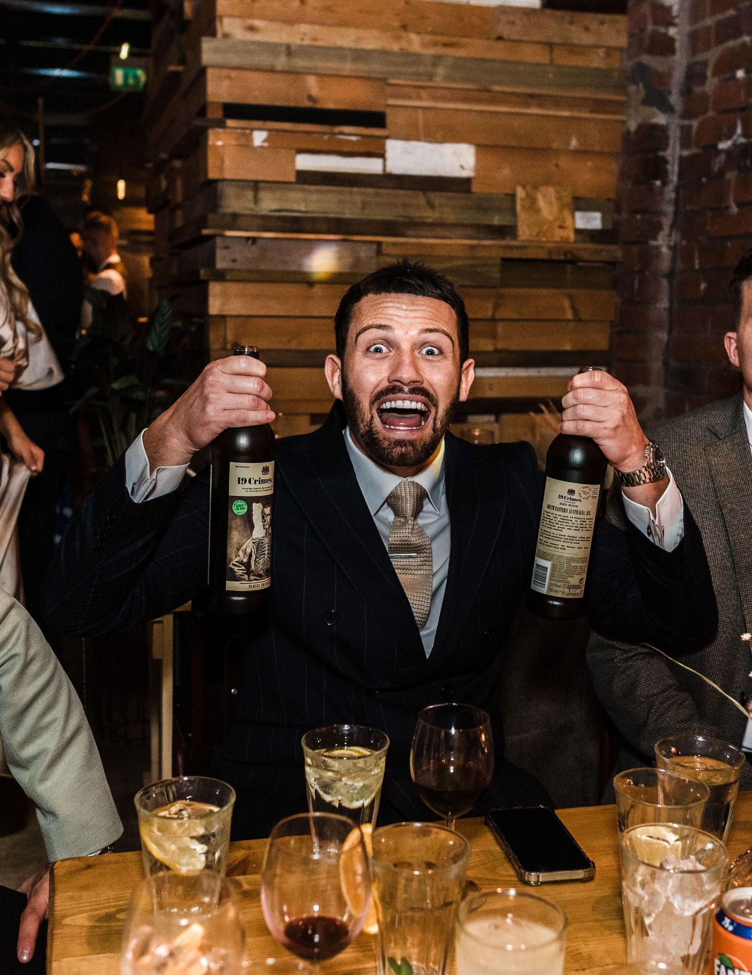 Enthusiastic party guest in suit holding up two bottles at rustic venue with wooden walls.