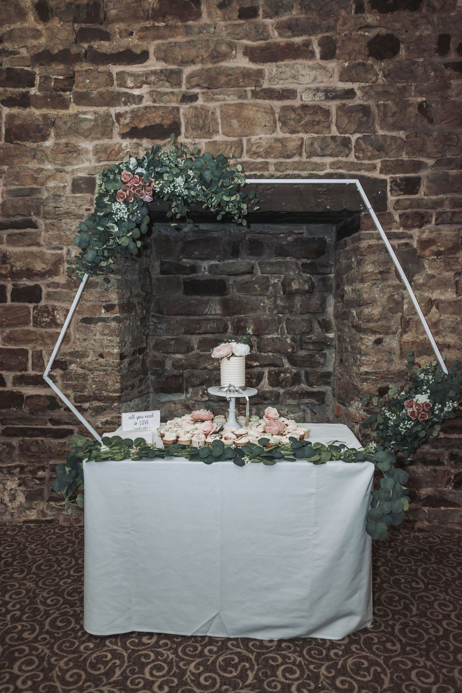 Wedding cake display table with geometric arch and floral decorations against a rustic brick wall.
