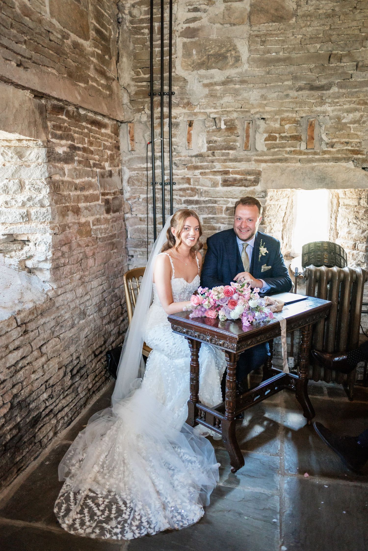 Newlyweds sit at rustic table with pink floral arrangements inside historic stone wall venue.