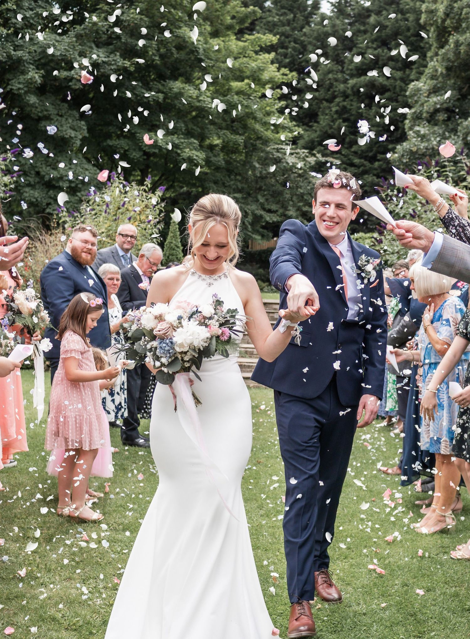 Wedding couple walking through confetti shower thrown by guests on garden lawn.