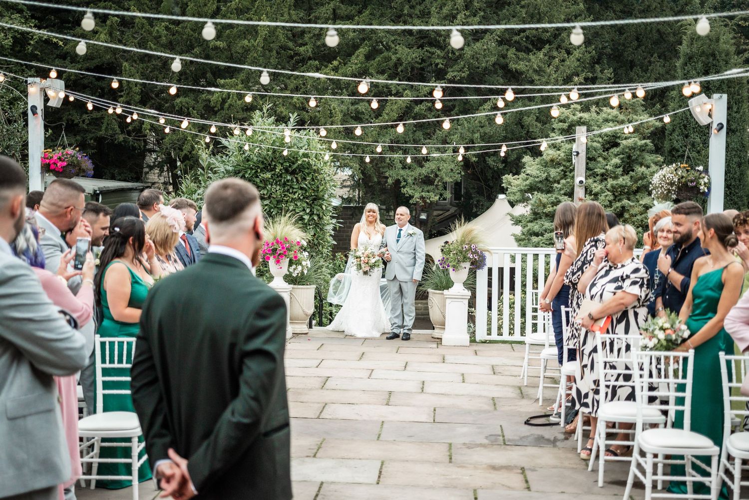 Outdoor wedding ceremony with string lights and white chairs on stone patio.