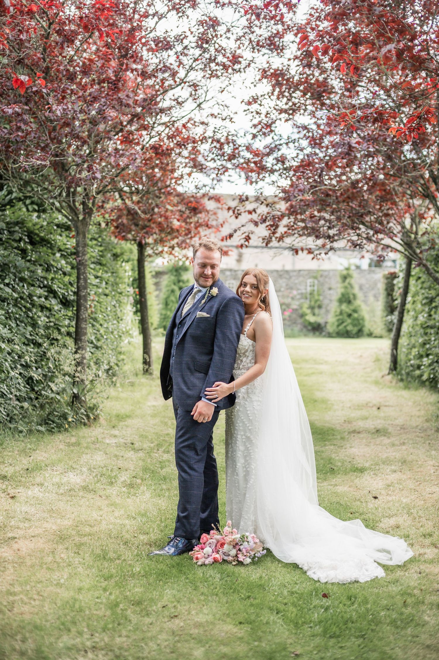 Elegant wedding couple stands in garden path framed by red trees and hedges.